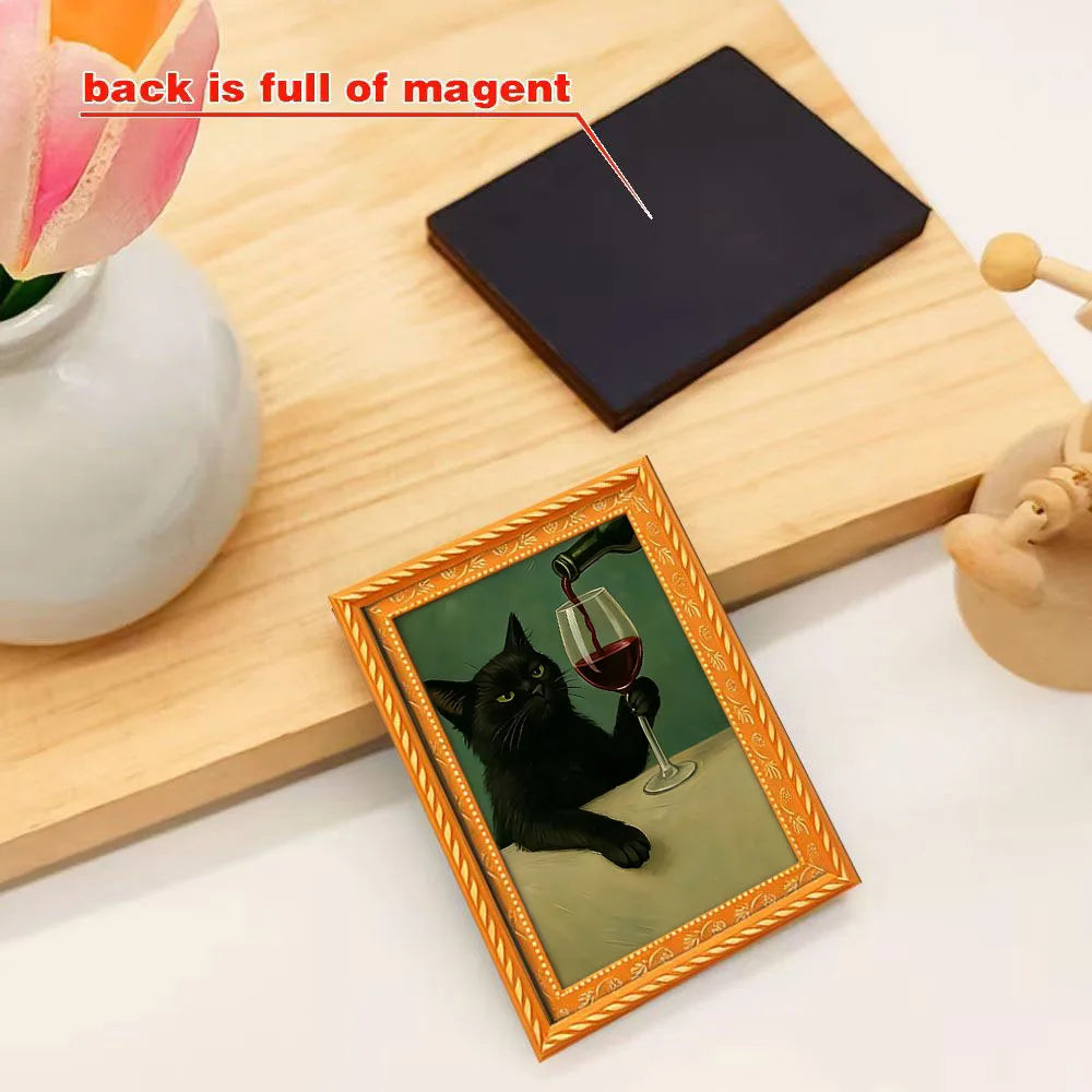 A framed fridge magnet featuring a black cat drinking from a glass of red wine, set on a wooden board.
