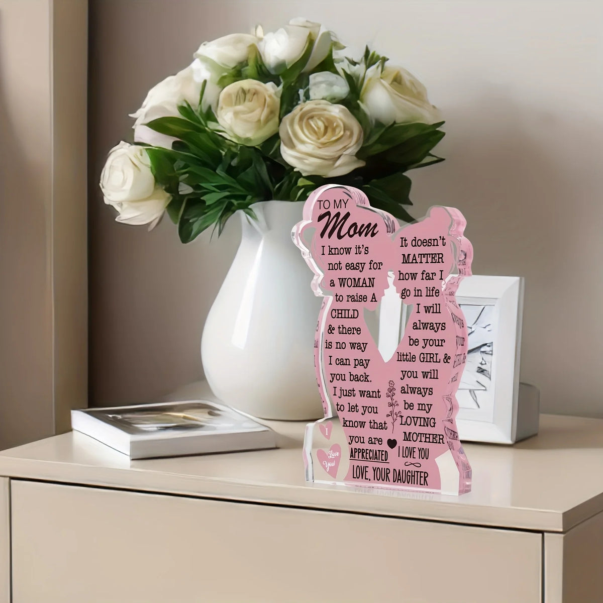 A white ceramic vase holds a bouquet of white roses and a personalized acrylic plaque for Mother's Day gifts.
