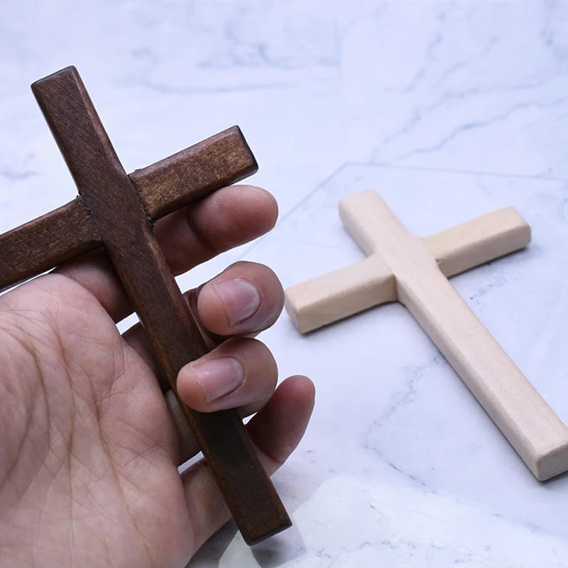 A hand holds a dark brown wooden wall cross and a light beige wooden wall cross on a white marble surface.