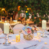 A table setting featuring a glass jar of flowers and several pillar candles on a white tablecloth.