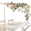 A gold stand holds a floral balloon arch decoration featuring white flowers, greenery, and gold accents above a wooden table.