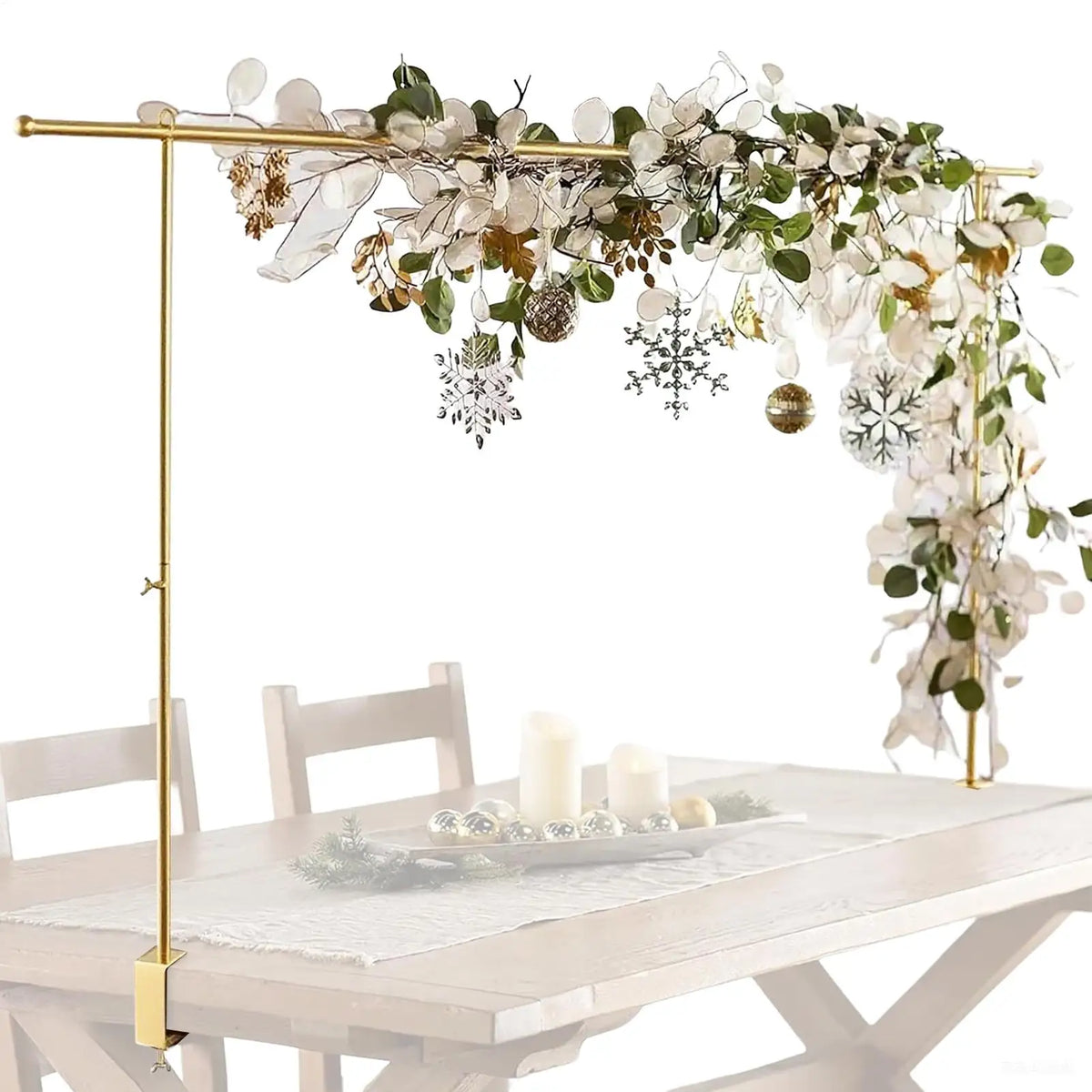 A gold stand holds a floral balloon arch decoration featuring white flowers, greenery, and gold accents above a wooden table.