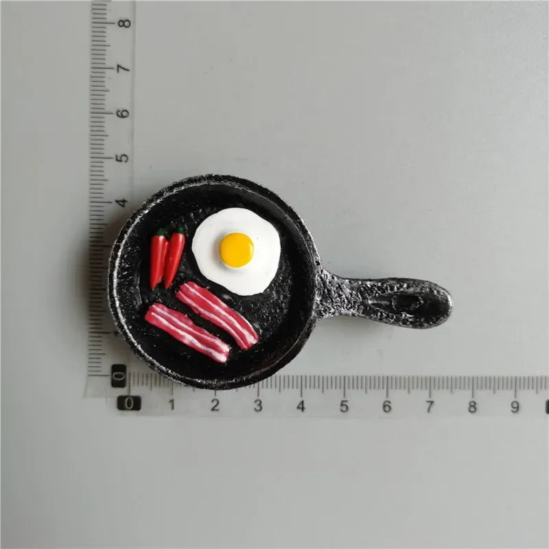 A black frying pan contains food-shaped fridge magnets including a fried egg and sausages.