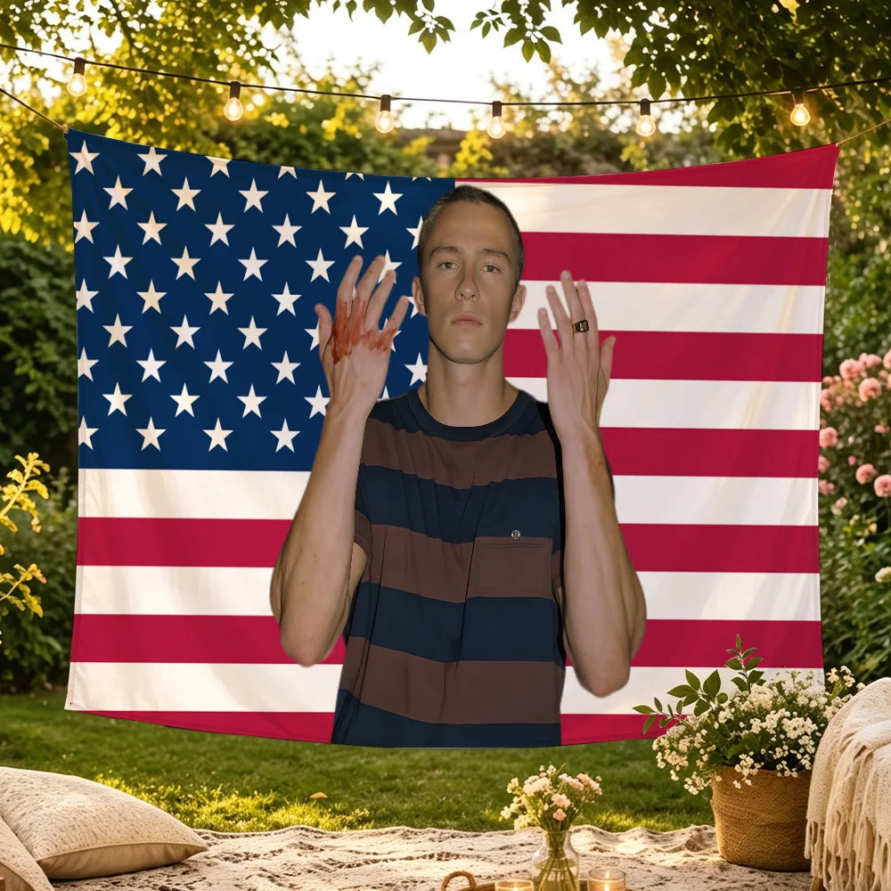 A young man stands in front of an American flag backdrop with a string of lights, holding his hands up.
