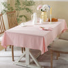 A pink classic cotton rectangle tablecloth is set on a wooden dining table with floral greenery in the background.