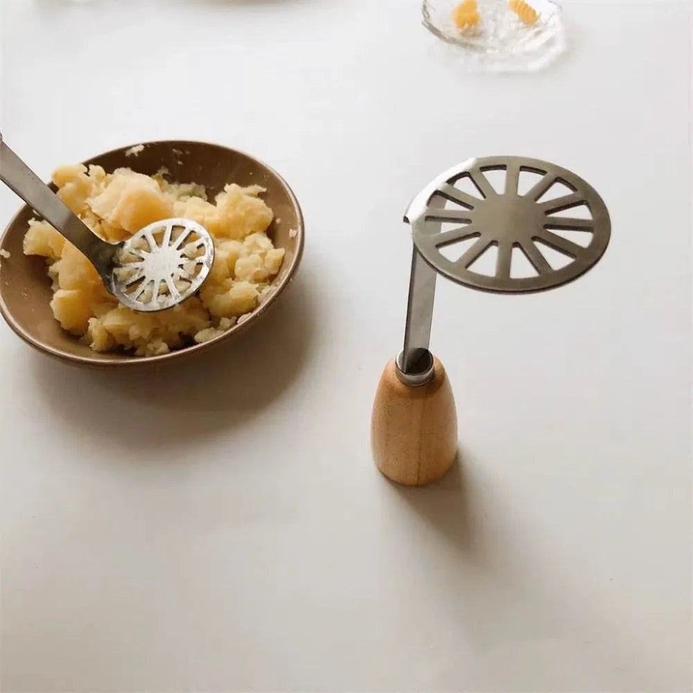 A manual wooden potato and pumpkin masher is shown next to a bowl of mashed food on a white surface.