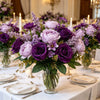 A lush bouquet of artificial mixed flowers featuring purple and pink roses, daisies, and lavender in a glass vase on a white 