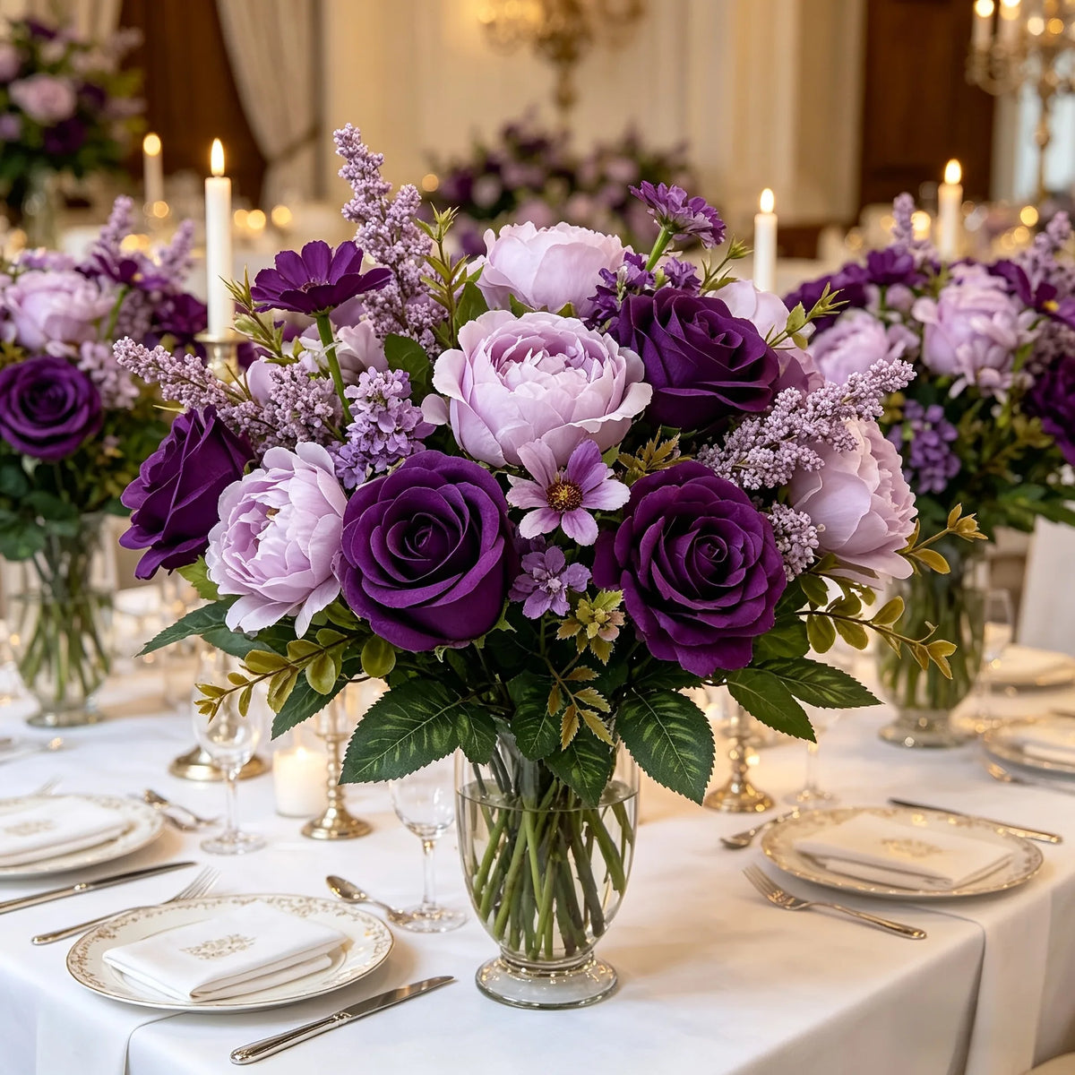 A lush bouquet of artificial mixed flowers featuring purple and pink roses, daisies, and lavender in a glass vase on a white 
