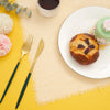 A set of ten handcrafted fringed cotton napkins displayed on a light beige linen cloth with a tea cup and a baked good.