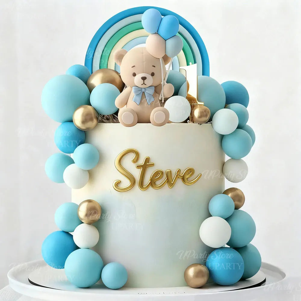 A white cake decorated with blue and gold balloons, featuring a teddy bear topper and a rainbow arch.
