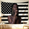 A person with dreadlocks is looking down, touching their face, positioned in front of a black and white American flag backdro