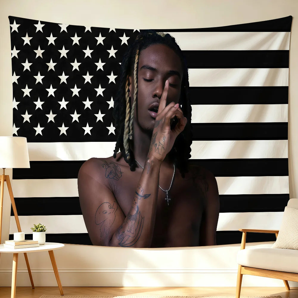 A person with dreadlocks is looking down, touching their face, positioned in front of a black and white American flag backdro