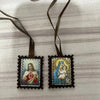 A pair of black wool scapulars with embroidered images of Jesus and Mary, tied with brown fabric ribbons.