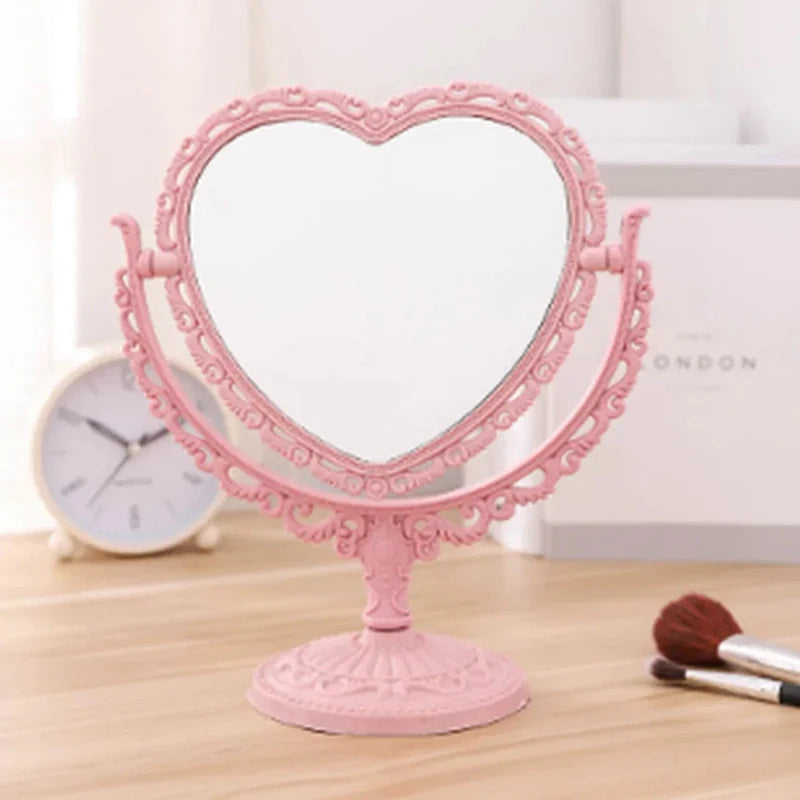 Espejo de maquillaje con forma de corazón rosa y marco ornamentado sobre una superficie de madera.
