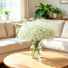 A clear glass vase filled with white baby's breath flowers sits on a wooden table next to a light-colored sofa.