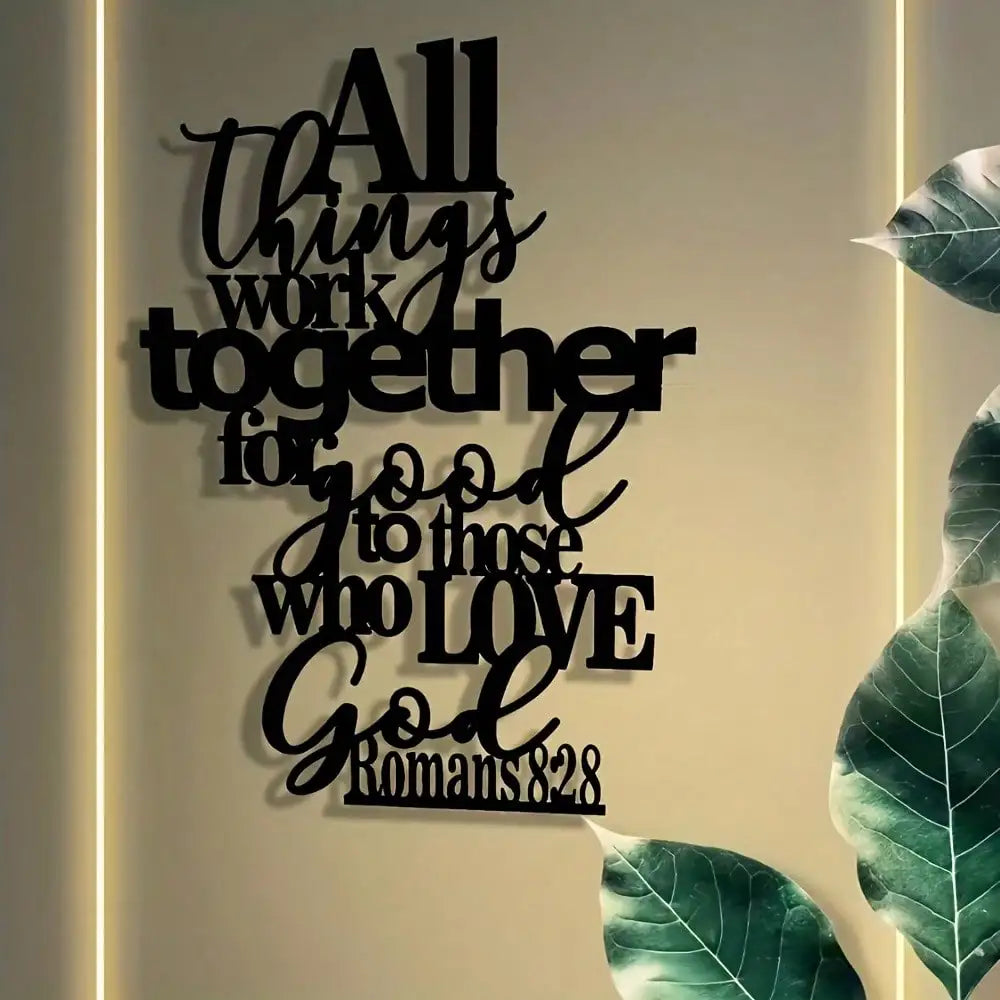 Black metal wall art featuring the Bible verse Romans 8:28 in a script font on a light beige wall.