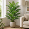 A tall artificial areca palm tree in a beige pot sits on a wooden floor next to a light-colored sofa.