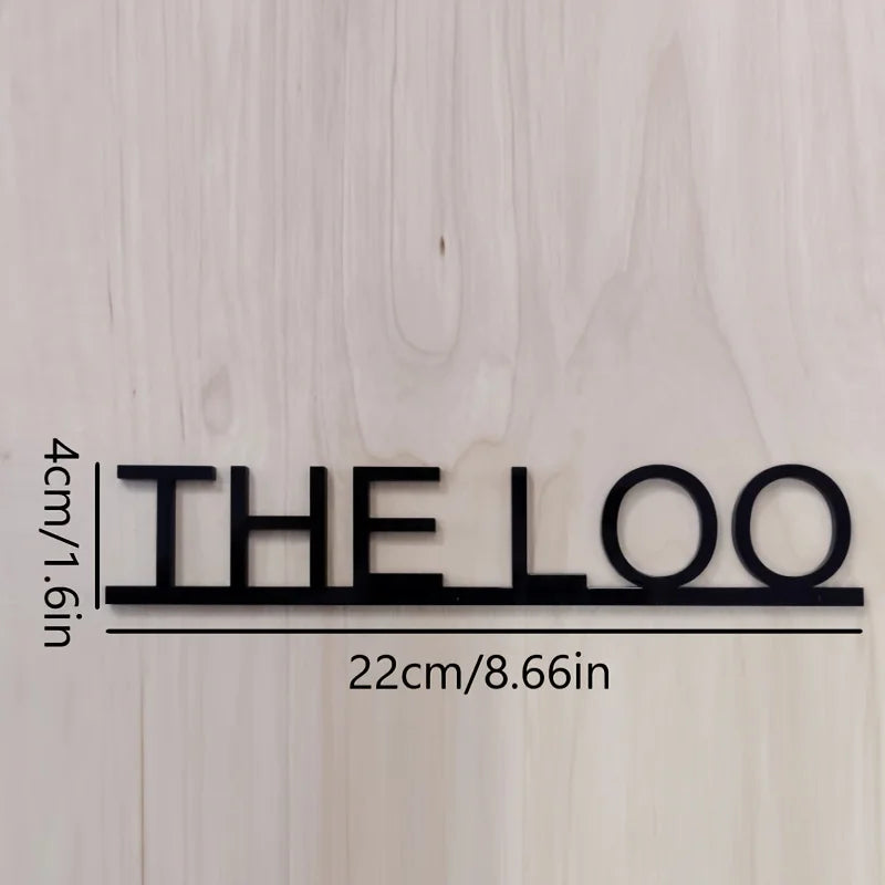 Un letrero de metal negro que deletrea THE LOO está montado en una pared de madera clara, con dimensiones indicadas.