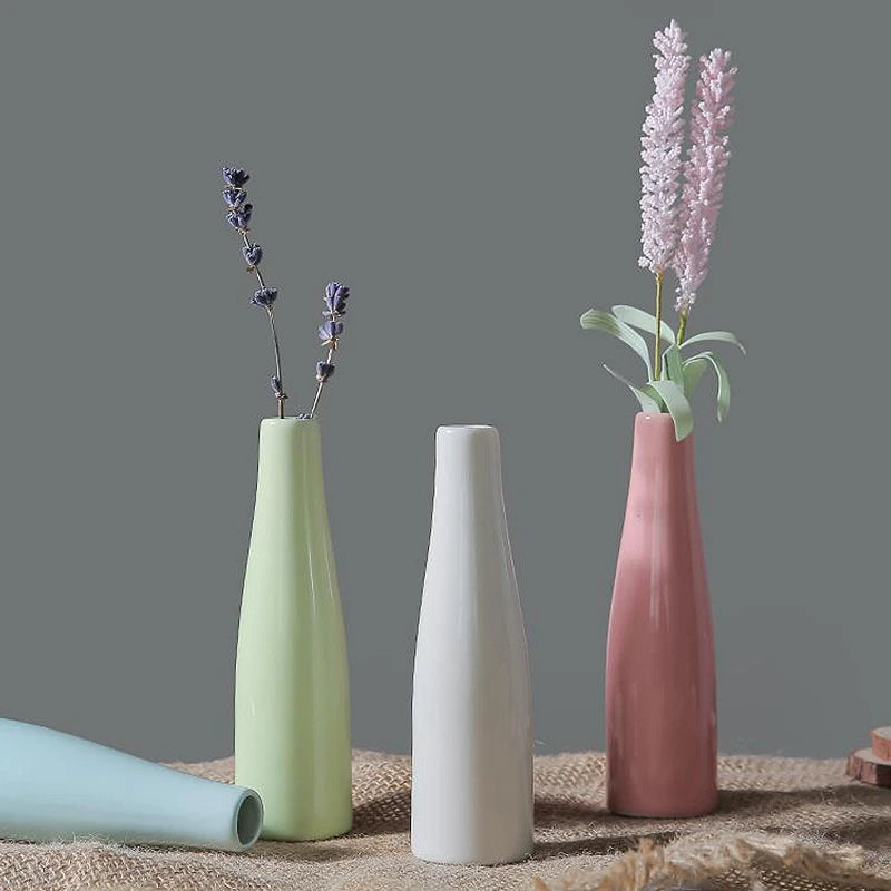 A pink ceramic vase with purple flowers stands on a woven fabric next to a light green vase.