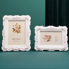 A white ornate frame with a floral design and a cream background is displayed on a teal surface.