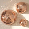 Trois miroirs ronds, boules de verre rose gold, suspendus au mur projetant des motifs lumineux sur une surface blanche.