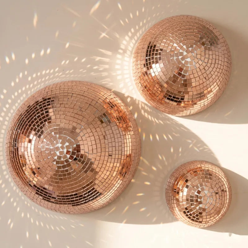 Trois miroirs ronds, boules de verre rose gold, suspendus au mur projetant des motifs lumineux sur une surface blanche.