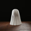 A white ghost figurine with a smooth, draped shape is displayed on a dark wooden surface against a black background.
