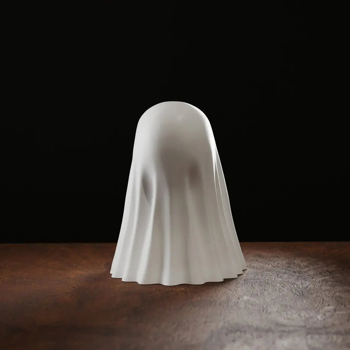 A white ghost figurine with a smooth, draped shape is displayed on a dark wooden surface against a black background.