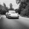 Une Porsche 911 argentée roule sur une route entourée d'arbres en noir et blanc.