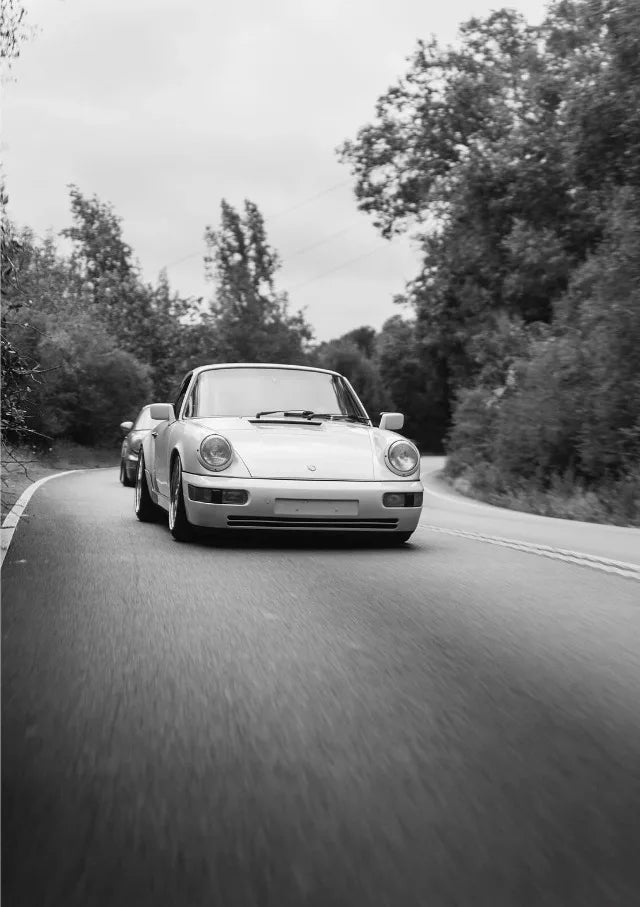 Une Porsche 911 argentée roule sur une route entourée d'arbres en noir et blanc.