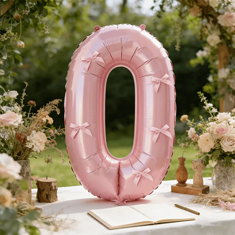 A large pink foil number balloon shaped like zero sitting on a white table surrounded by floral greenery.