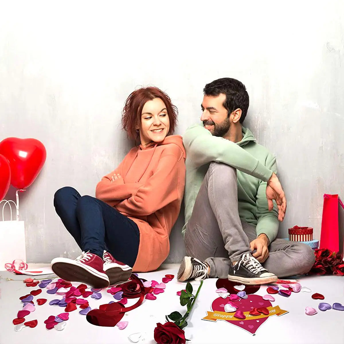 A couple sits on a white surface surrounded by red and pink heart petals, with a red heart balloon in the background.