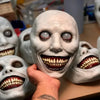 A hand holds a realistic smiling demon latex mask with white skin and visible teeth.