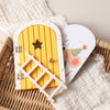 A wooden fairy elf door miniature decoration with a tooth box, featuring a yellow arched door and a white picket fence.