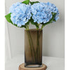 A bouquet of light blue artificial hydrangea flowers in a tall glass vase on a wooden coaster.