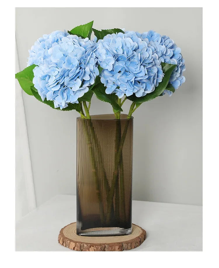A bouquet of light blue artificial hydrangea flowers in a tall glass vase on a wooden coaster.