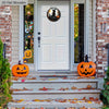Porte blanche avec une enseigne ronde d'Halloween représentant une sorcière et un chat, flanquée de feuilles d'automne.
