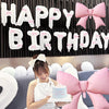 A woman in a white dress holds a cake and is positioned in front of a black banner decorated with white letters and pink bows