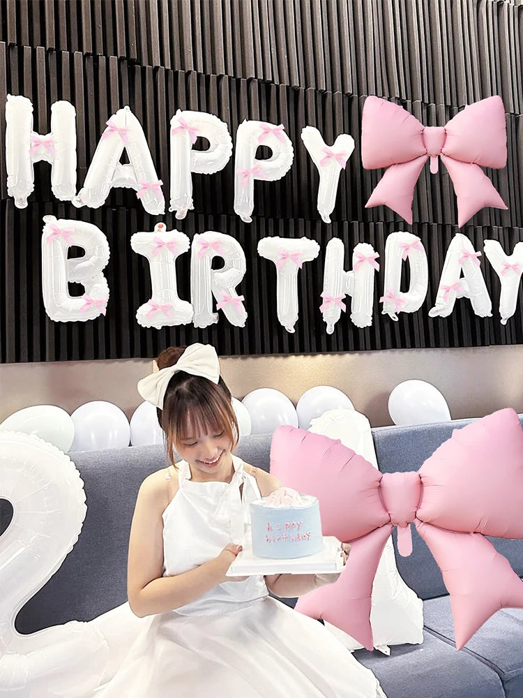 A woman in a white dress holds a cake and is positioned in front of a black banner decorated with white letters and pink bows