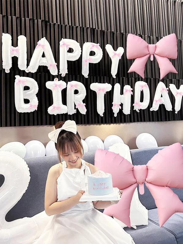 A woman in a white dress holds a cake and is positioned in front of a black banner decorated with white letters and pink bows
