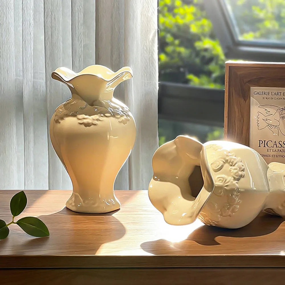 A light beige ceramic vase with floral relief detailing sits on a wooden surface next to a green leaf.