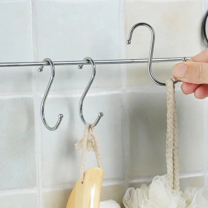 A hand is hanging a beige fabric loop onto a set of stainless steel S-shape hanging hooks mounted on a tiled wall.