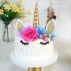 A white unicorn cake decorated with pink roses, blue flowers, gold horn, and glitter accents.