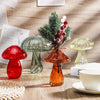 A still life featuring three mushroom-shaped glass vases in translucent red, green, and amber tones on a wooden surface.