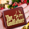 A hand holds a brown music box engraved with the text The Godfather against a festive green Christmas tree background.