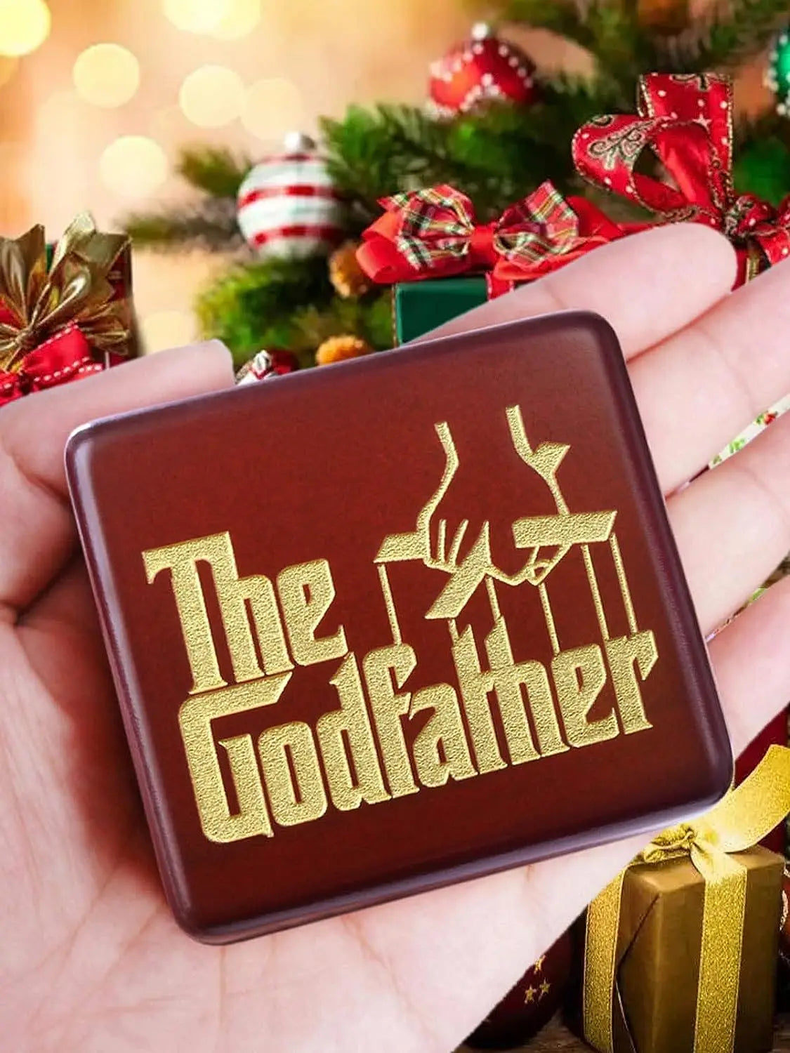 A hand holds a brown music box engraved with the text The Godfather against a festive green Christmas tree background.