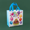 A white reusable happy birthday gift bag with blue handles decorated with colorful balloons and confetti.