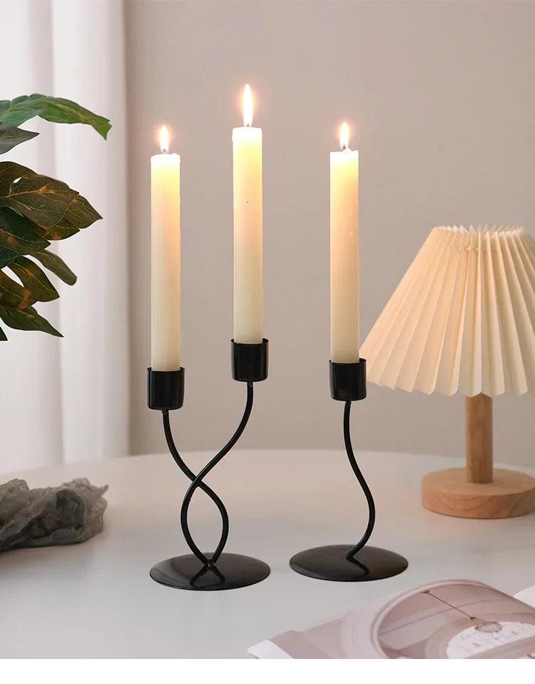 A set of black iron candle holders with white taper candles on a white table next to a cream-colored lampshade.