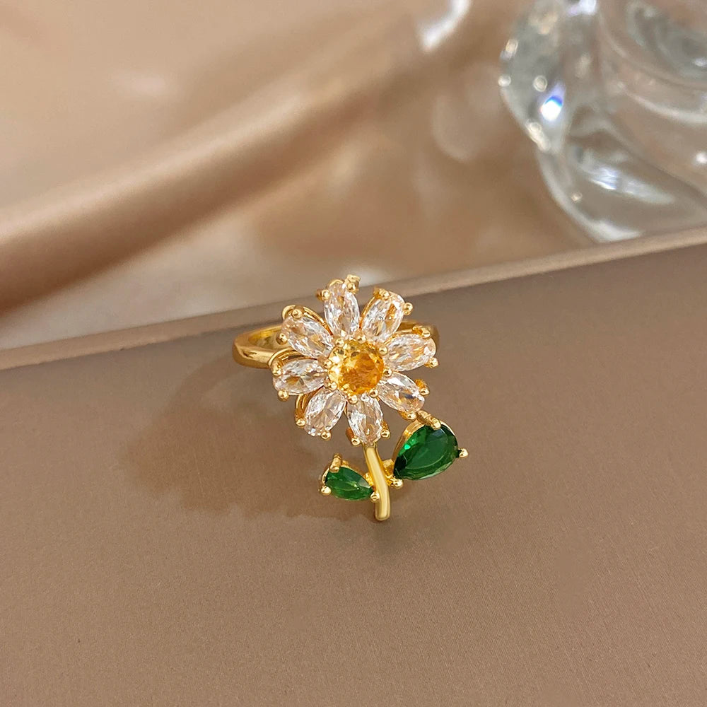 A gold cocktail ring featuring a flower design with clear stones and an attached green leaf.