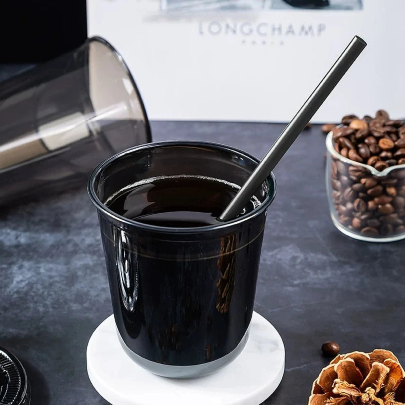 Eine schwarze Einwegtasse mit Strohhalm, gefüllt mit dunkler Flüssigkeit, steht auf einer weißen Untertasse.